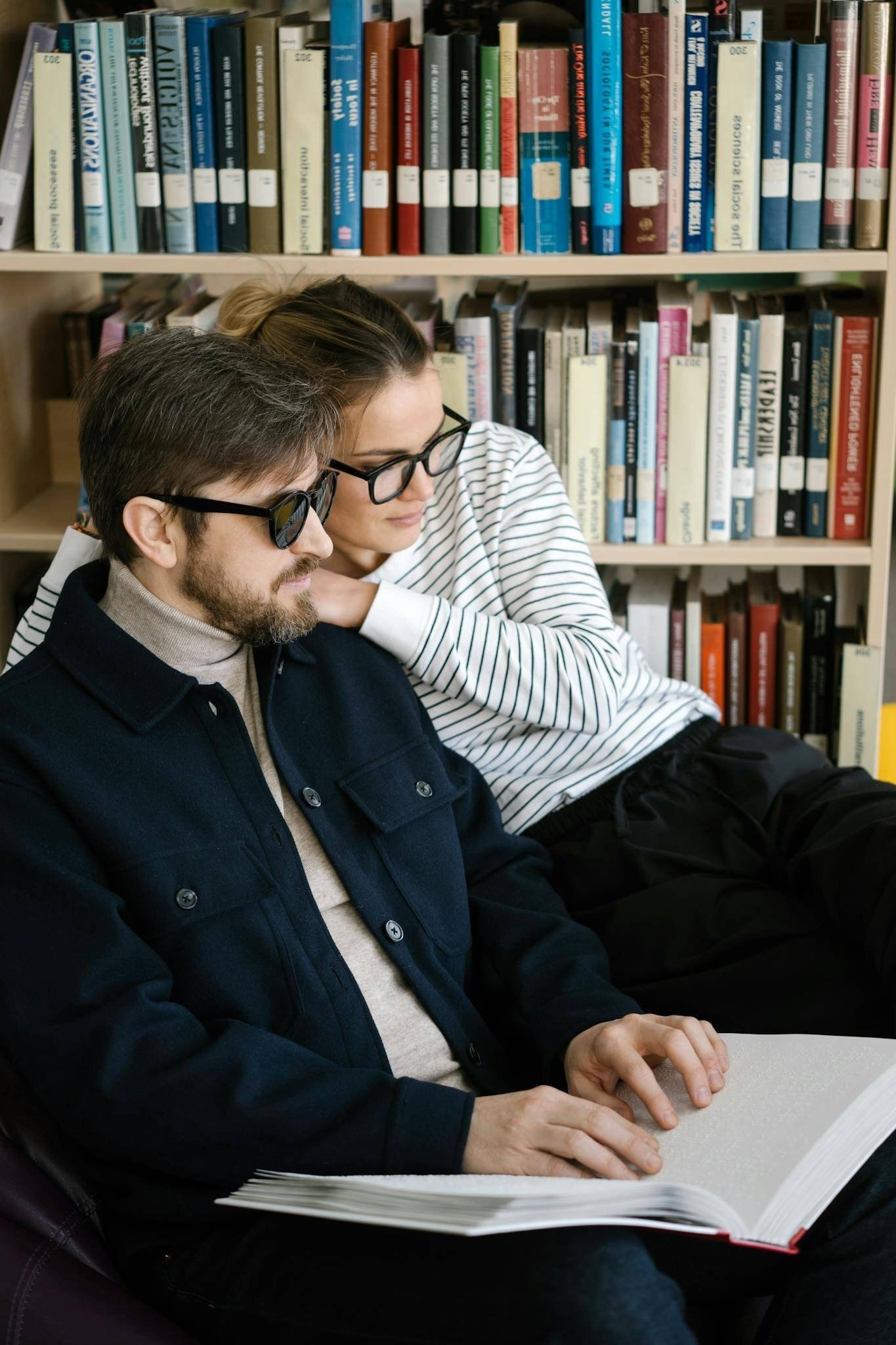 A couple reading a large book together in a library, surrounded by shelves of books.